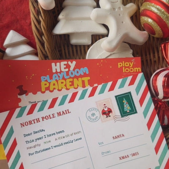 Child's hand painting a ceramic gingerbread man from a Playloom Christmas craft kit, with a festive fireplace and Christmas tree in the background.