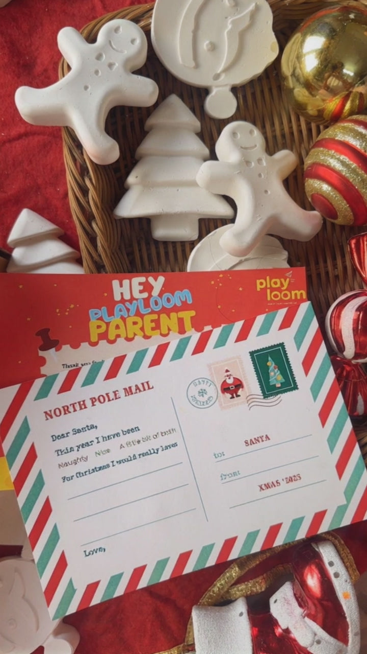 Child's hand painting a ceramic gingerbread man from a Playloom Christmas craft kit, with a festive fireplace and Christmas tree in the background.