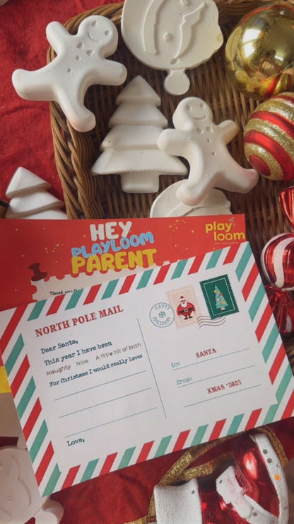 Child's hand painting a ceramic gingerbread man from a Playloom Christmas craft kit, with a festive fireplace and Christmas tree in the background.
