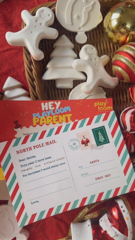 Child's hand painting a ceramic gingerbread man from a Playloom Christmas craft kit, with a festive fireplace and Christmas tree in the background.