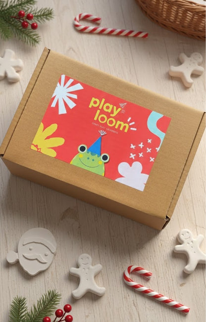 Stack of Playloom Christmas craft kit boxes ready for shipping, featuring the festive frog logo and surrounded by ceramic ornaments.